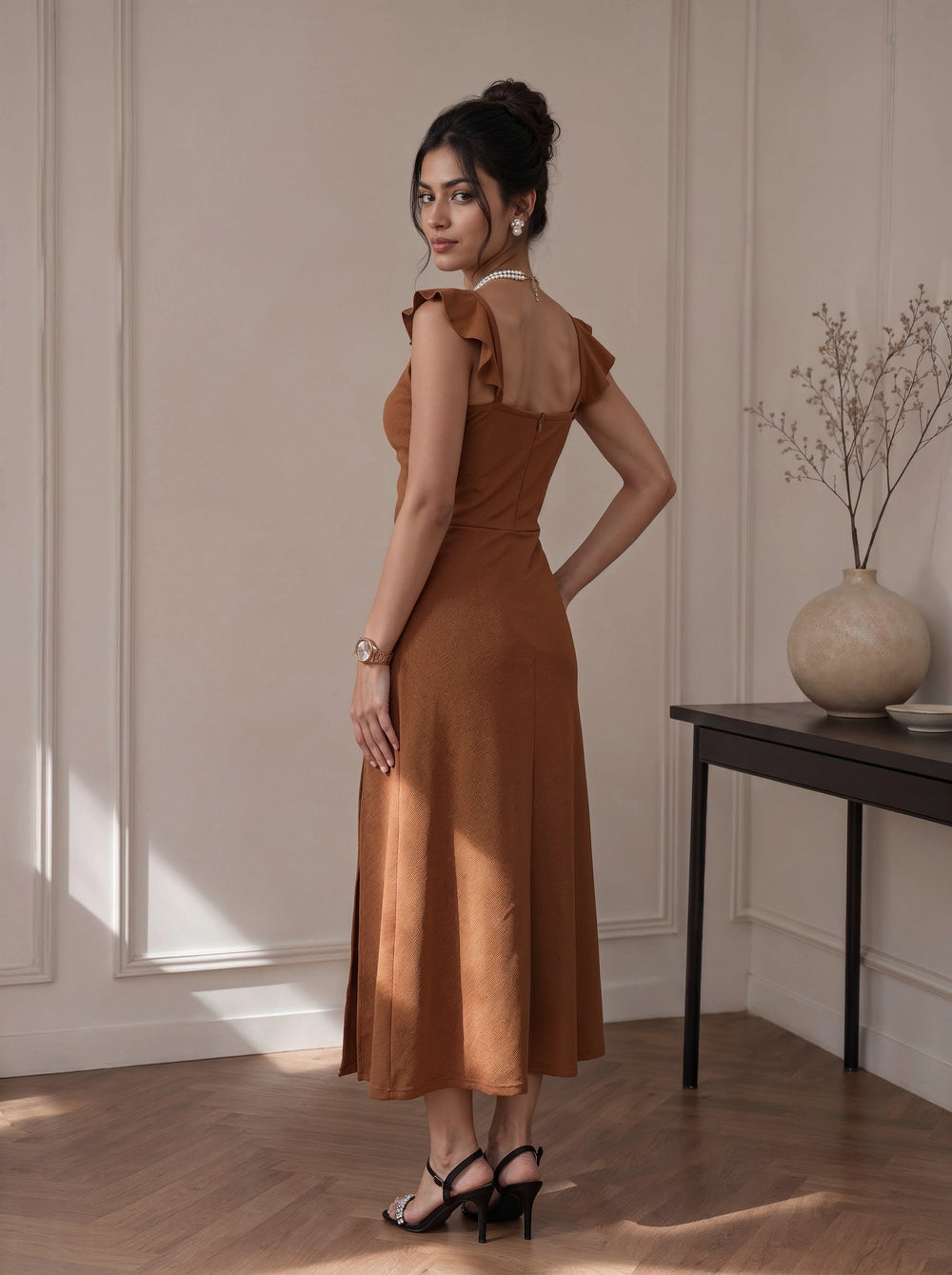 A woman stands in a sunlit room wearing a rust-brown sleeveless midi dress with ruffled cap sleeves and a sheer back panel, paired with black strappy heels.