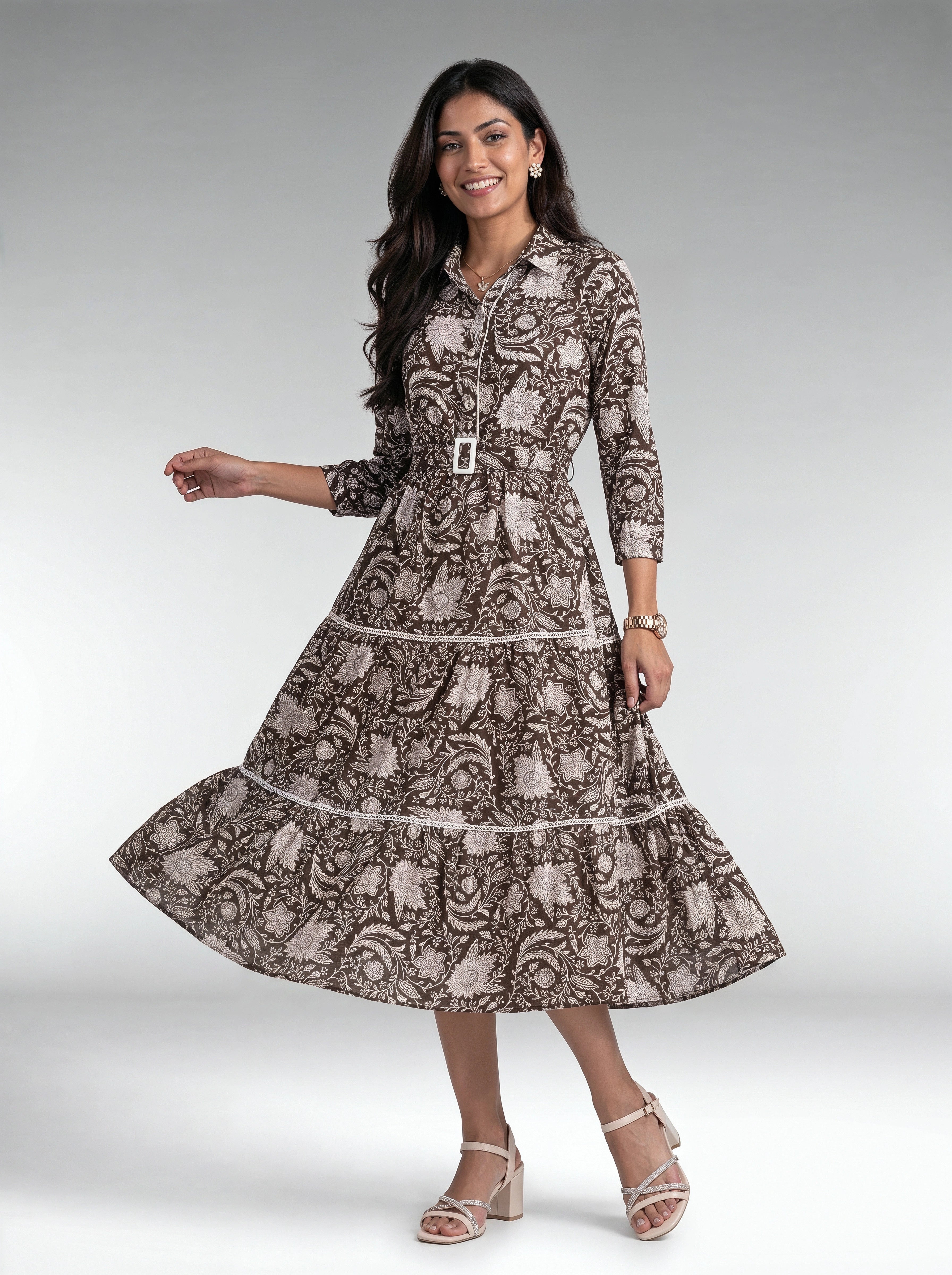 A woman modeling a brown and beige floral print midi dress with a collar, button-up front, and tiered skirt, paired with white heeled sandals against a gray studio background.