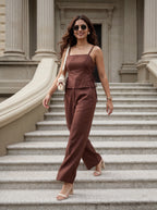 A woman wearing a brown sleeveless camisole and matching wide-leg trousers jumpsuit walks down grand stone columns and steps, carrying a small white shoulder bag.