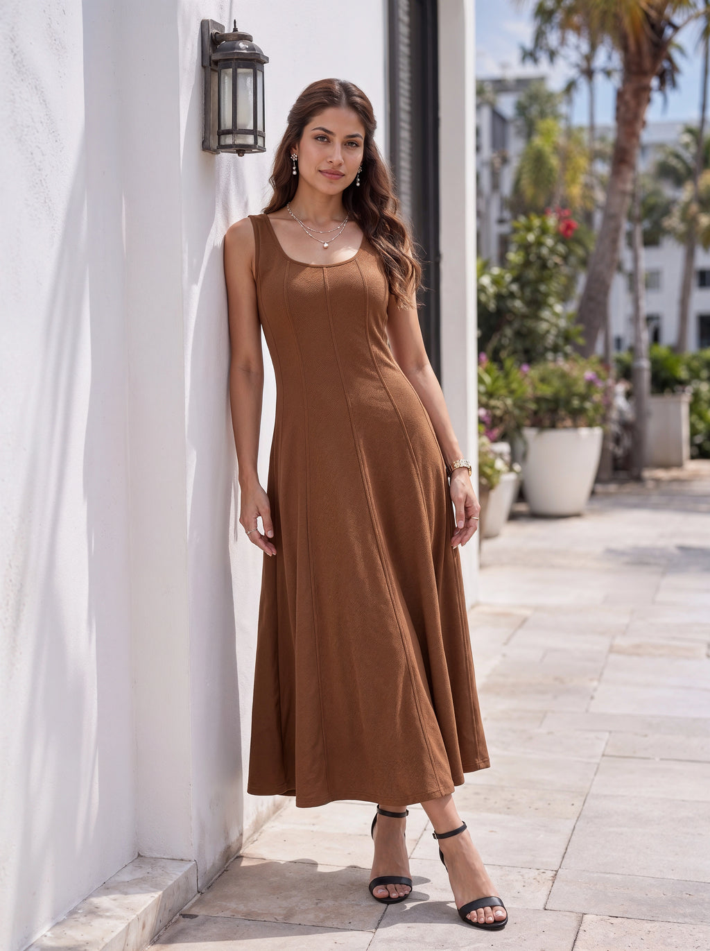 A woman wearing a sleeveless brown maxi dress and black strappy heels stands against a white stucco wall in a sunny outdoor setting with palm trees and potted plants in the background.