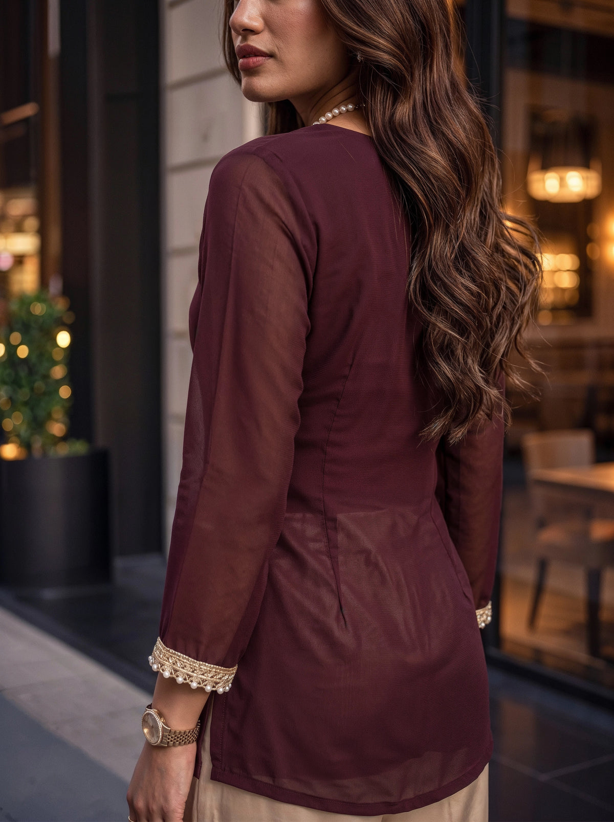 A woman shown from the back wearing a long-sleeved maroon tunic with gold lace trim at the cuffs, photographed in an outdoor urban setting.