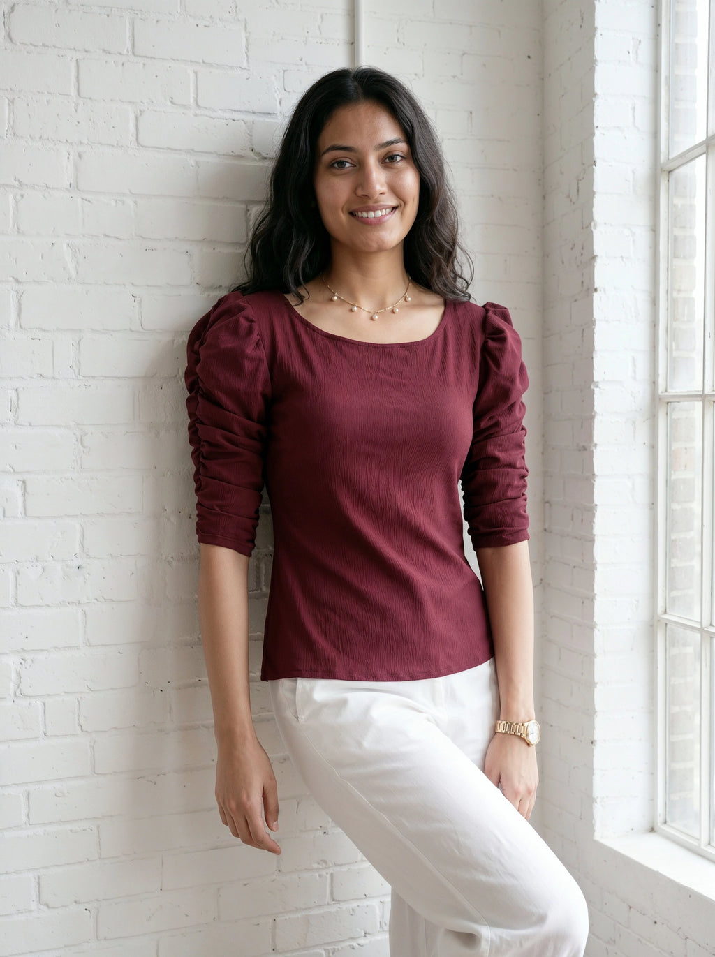 Woman wearing a burgundy top with puffed sleeves and white pants against a white brick wall.