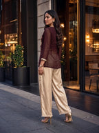 A woman wearing a dark brown long-sleeve kurta and beige wide-leg trousers with decorative trim, standing on a city sidewalk in front of a modern building.