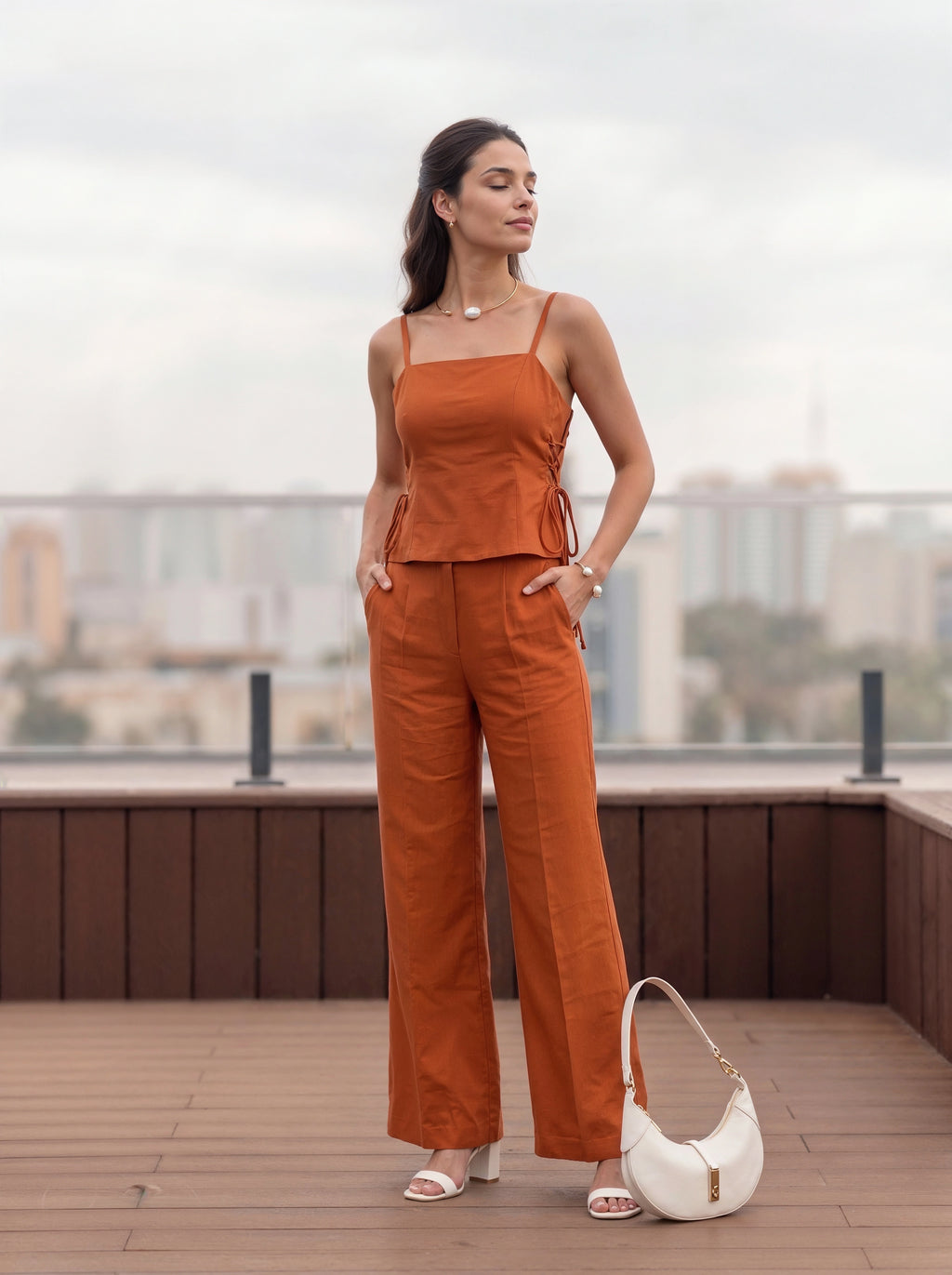 A woman stands on a rooftop terrace wearing a rust-orange sleeveless camisole and matching wide-leg pants, with a white handbag beside her. The background shows a blurred cityscape under an overcast sky.