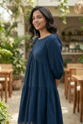 Woman in a blue dress standing outdoors with plants and tables in the background