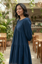 Woman in a blue dress standing outdoors with plants and tables in the background