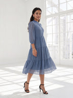 Woman wearing a blue dress standing in a bright room with large windows.
