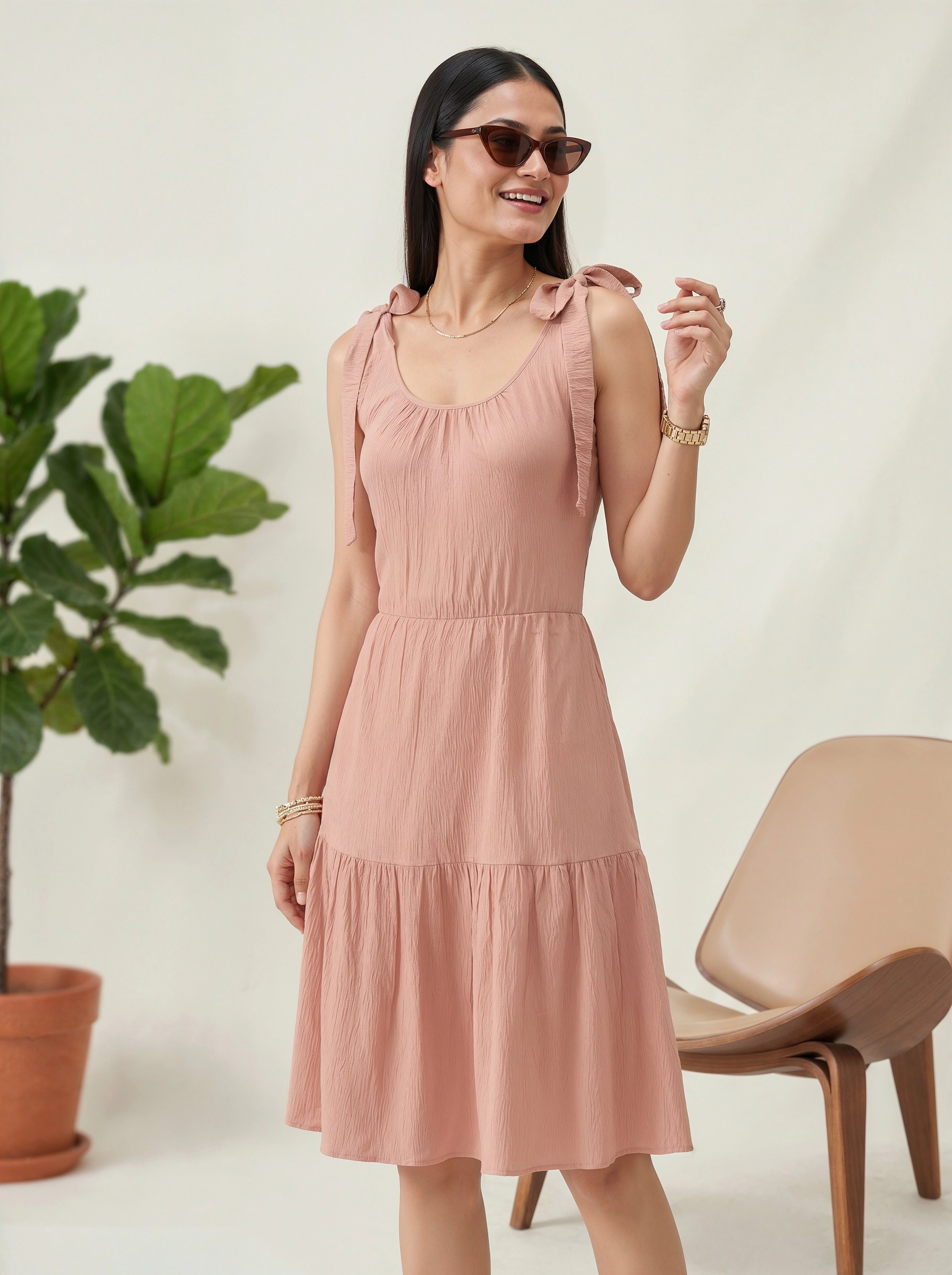 Woman wearing a pink dress standing next to a plant and chair against a plain background