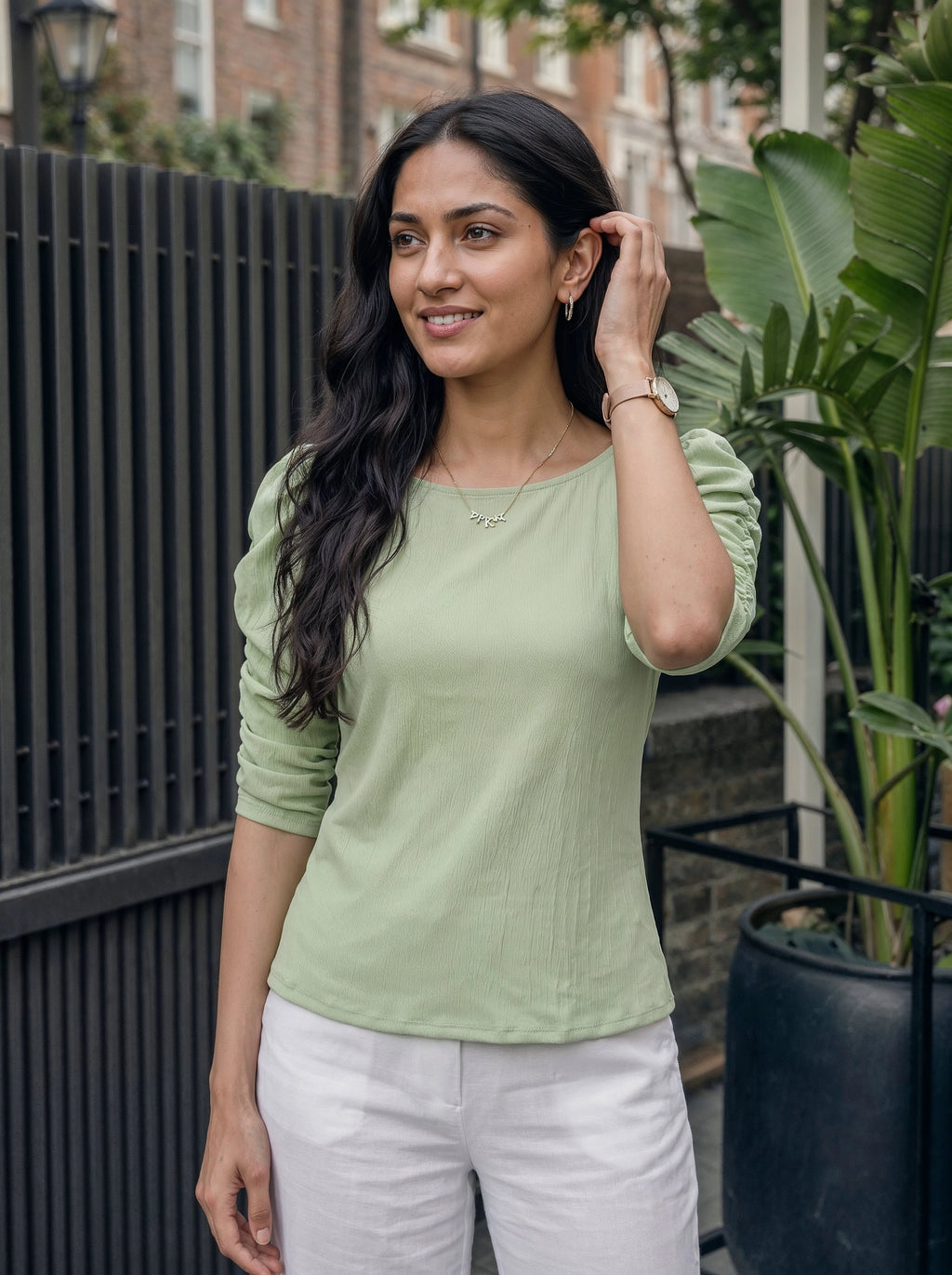 Woman wearing a green top and white pants standing outdoors with plants and a fence in the background