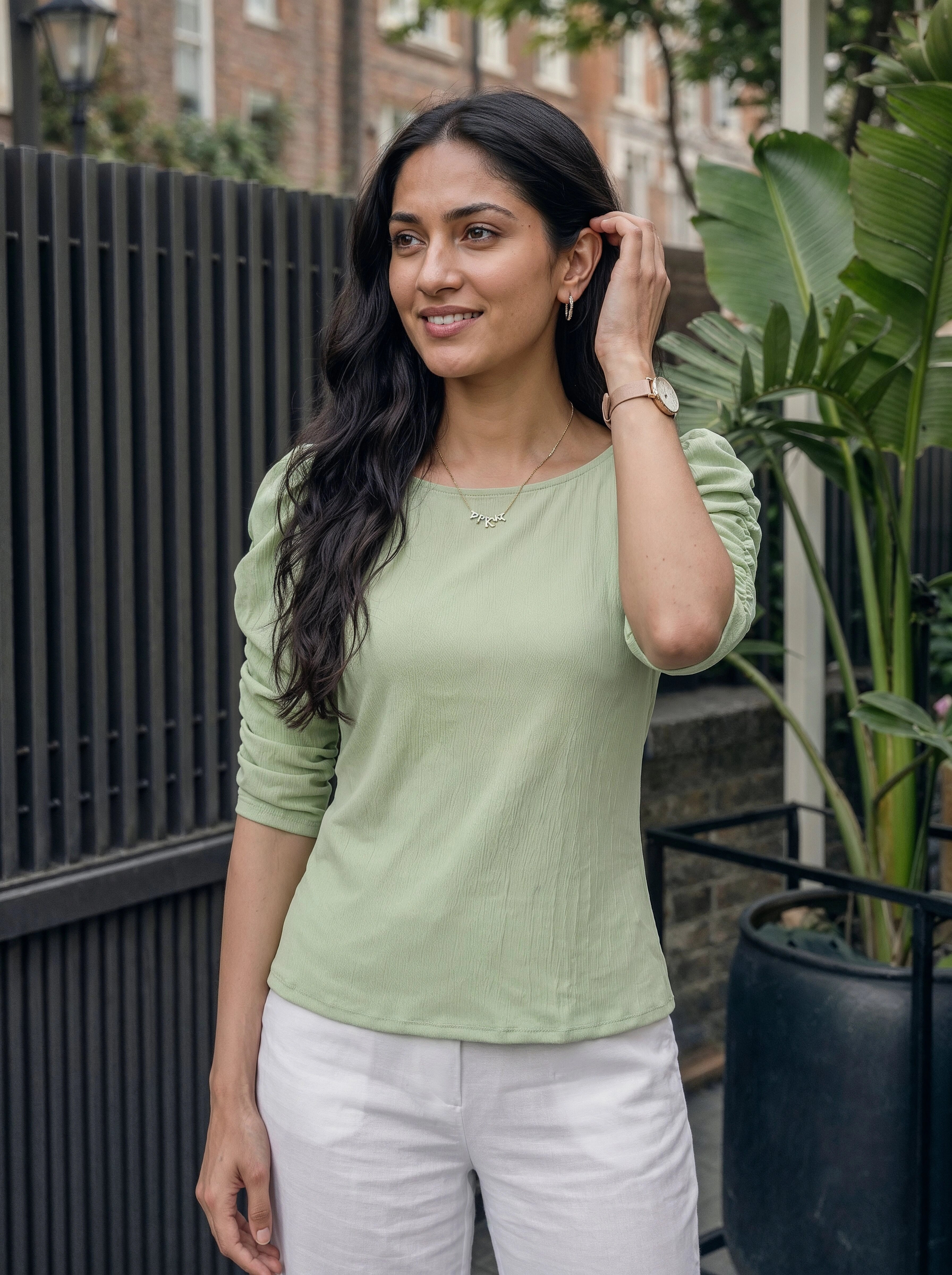Woman wearing a green top and white pants standing outdoors with plants and a fence in the background