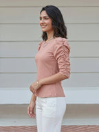 Woman wearing a pink blouse and white pants standing against a light-colored wall.