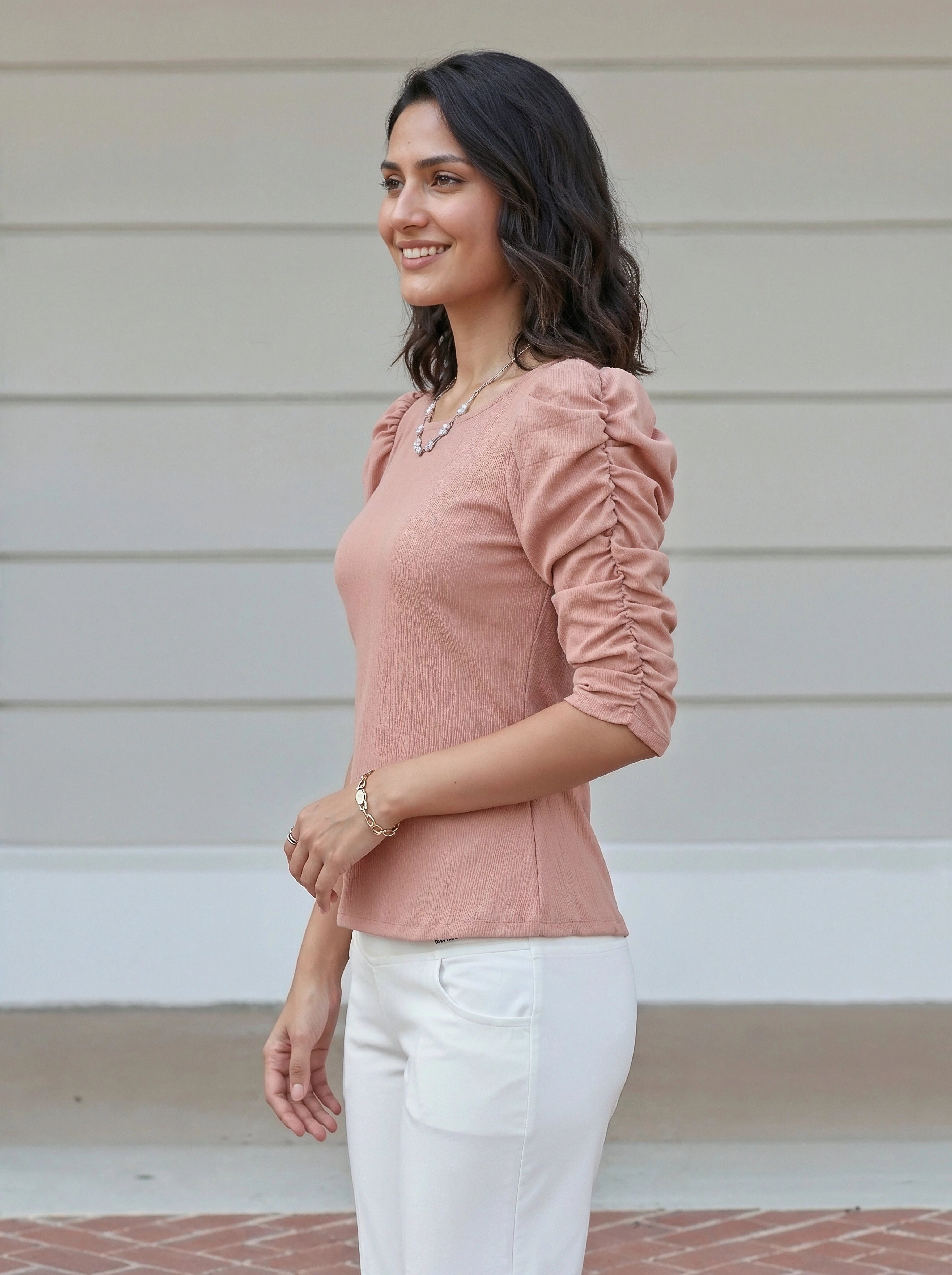 Woman wearing a pink blouse and white pants standing against a light-colored wall.