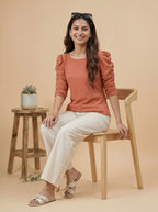 Woman sitting on a chair wearing a rust-colored top and white pants, with a wooden stool and plant in the background.