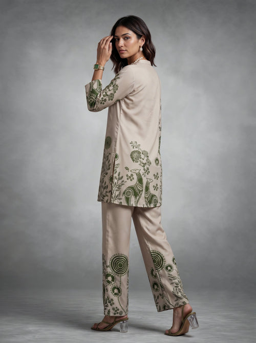A woman modeling a beige two-piece kurta and trouser set with green floral and paisley embroidery, posed against a neutral gray studio background.