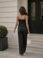 A woman viewed from behind wearing a black camisole-style top with a gold zipper at the back and matching wide-leg trousers, walking up stone steps outside a building and holding a small white handbag.