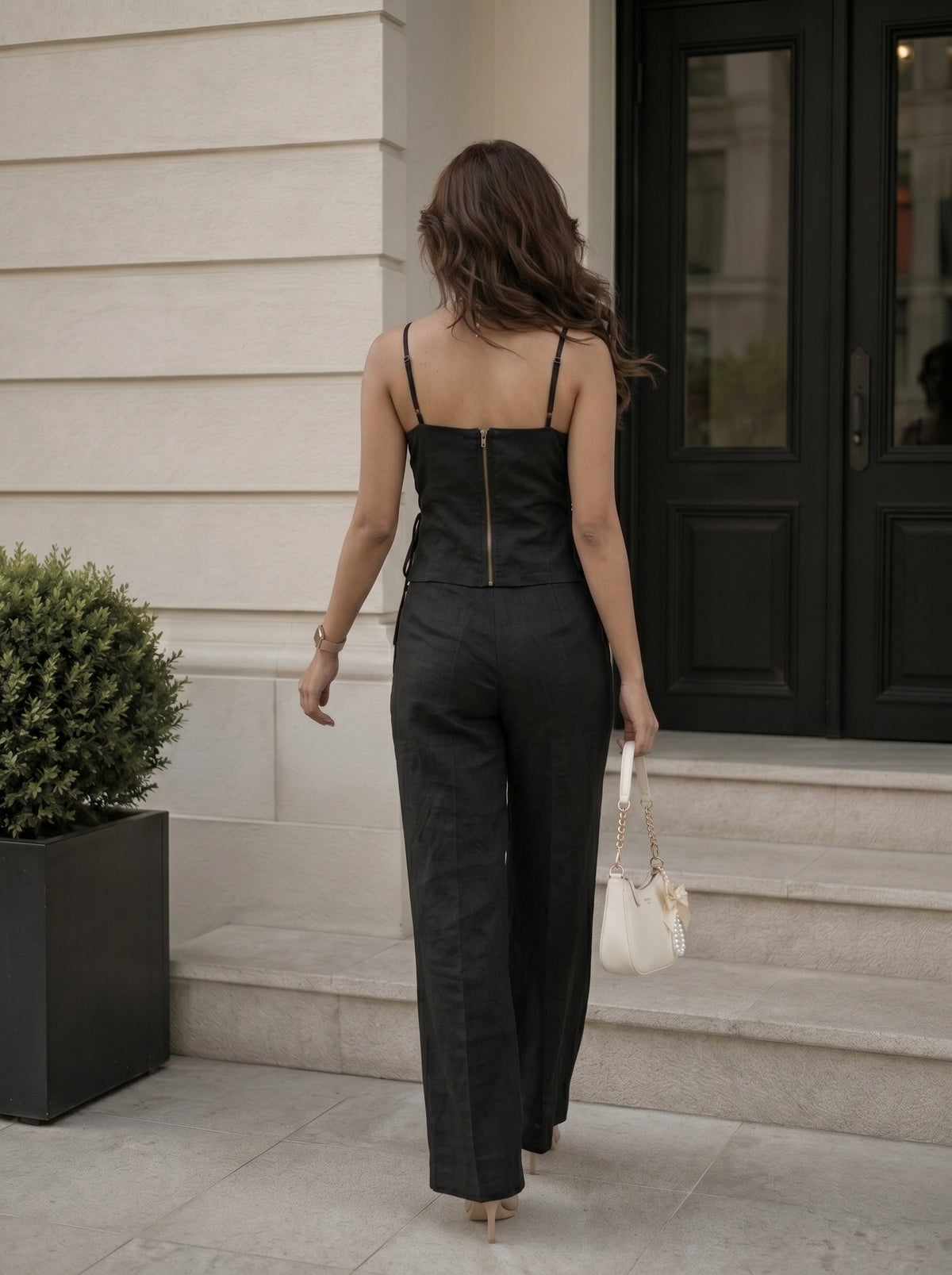 A woman viewed from behind wearing a black camisole-style top with a gold zipper at the back and matching wide-leg trousers, walking up stone steps outside a building and holding a small white handbag.