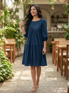 Woman in a blue dress standing in an outdoor setting with plants and tables.