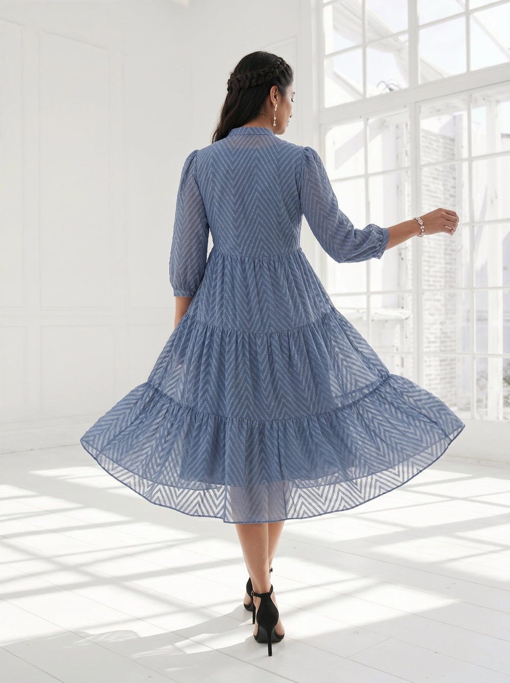 Woman in a blue dress standing in a bright room with large windows.