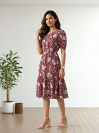A woman modeling a short-sleeve midi dress with a red and beige paisley pattern, standing in a bright room with a wooden floor and a potted plant.