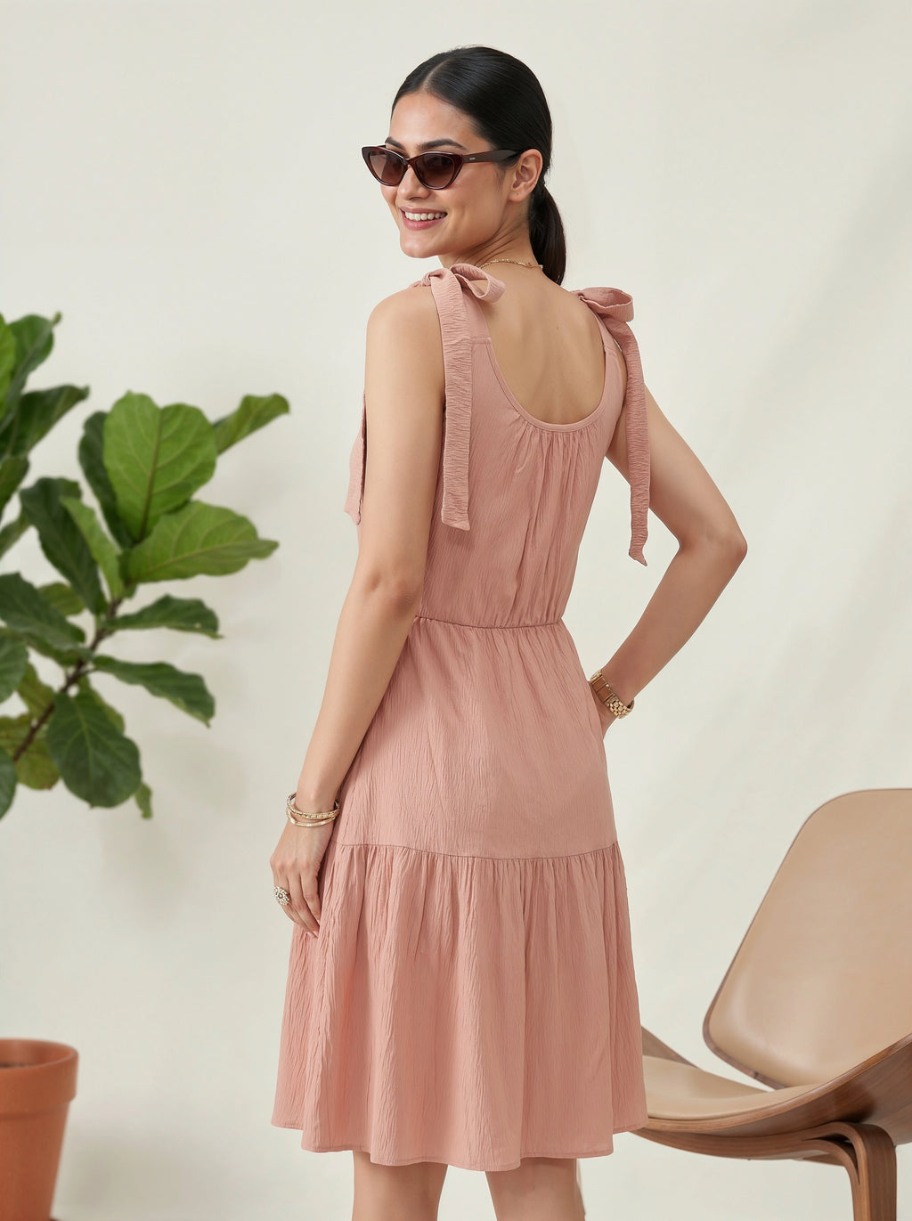 Woman wearing a pink dress with bow straps, standing against a white wall with a plant and chair in the background.