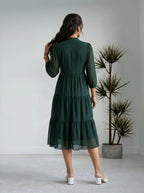 Woman wearing a green dress standing in a minimalistic room with a plant.
