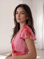 A woman with long brown hair wearing a pink short-sleeve dress with a red and black floral pattern, photographed in a bright, minimalist room with white curtains.