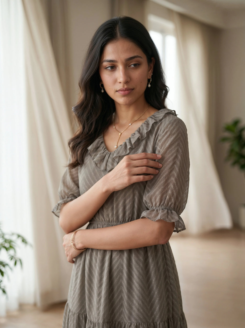 Woman wearing a gray dress standing in a softly lit room with curtains and plants.