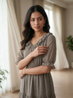 Woman wearing a gray dress standing in a softly lit room with curtains and plants.