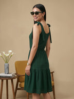 Woman in a green dress standing in a room with a beige wall, wooden chair, and small table with a vase.