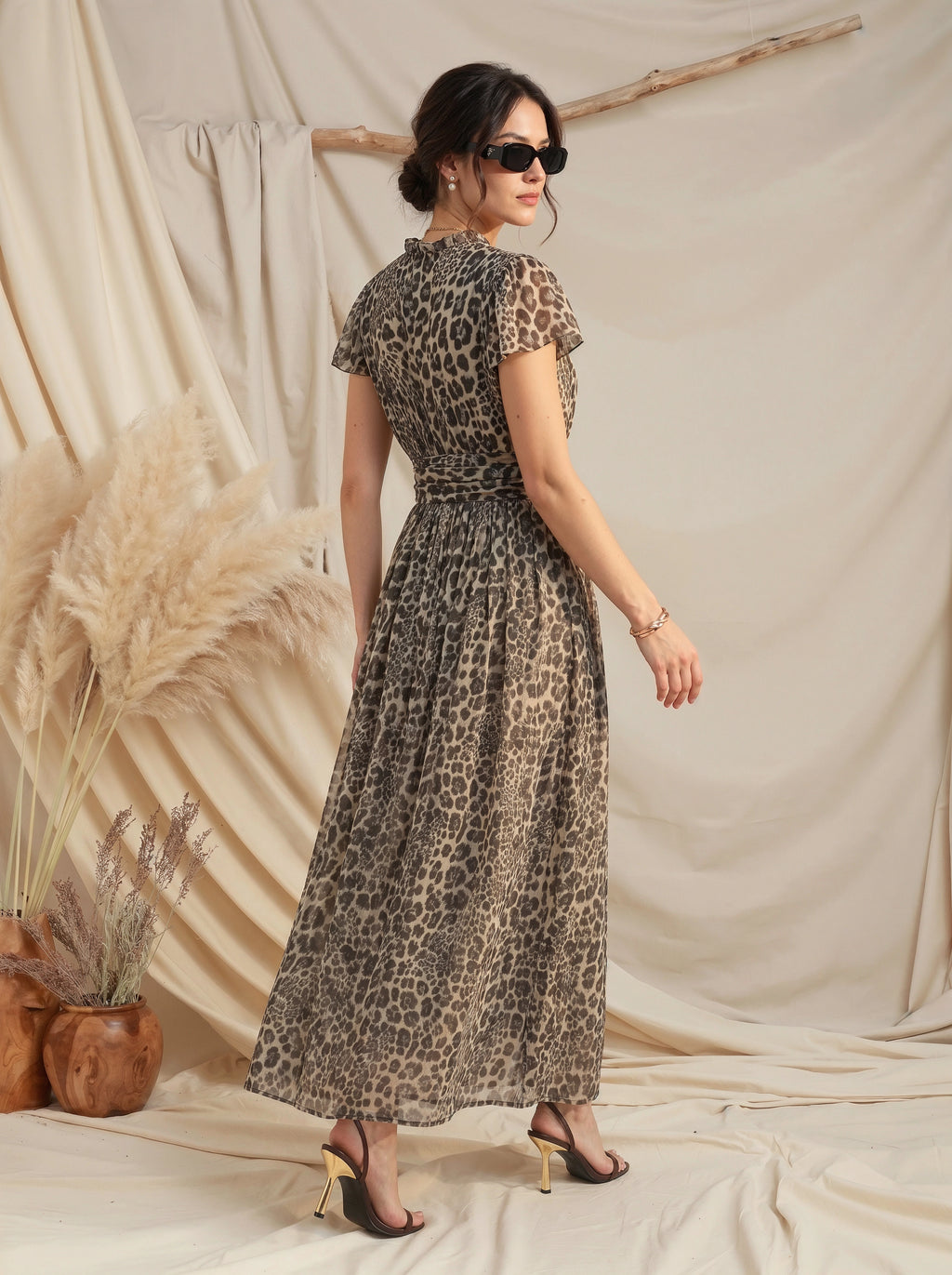 A woman modeling a leopard-print maxi dress with short flutter sleeves and a tie waist, photographed in a styled studio setting with draped beige fabric, pampas grass, and wooden decor.