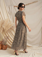 A woman modeling a leopard-print maxi dress with short flutter sleeves and a tie waist, photographed in a styled studio setting with draped beige fabric, pampas grass, and wooden decor.