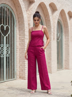 A woman wearing a magenta two-piece jumpsuit with a peplum top and wide-leg pants, standing outdoors in front of a beige brick building with arched windows.