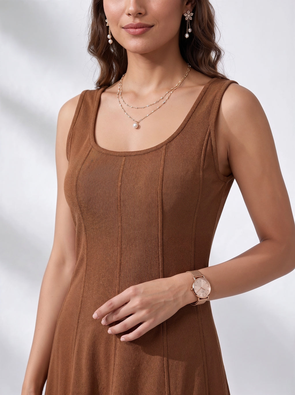 A woman wearing a sleeveless brown ribbed knit dress, styled with layered gold necklaces, dangling earrings, and a rose gold watch, set against a softly lit white background.