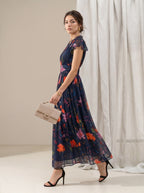 A woman models a navy blue maxi dress with a vibrant orange, pink, and red floral print. She is holding a beige clutch bag and wearing black high-heeled sandals, posed in front of a neutral beige wall and white draped fabric backdrop.