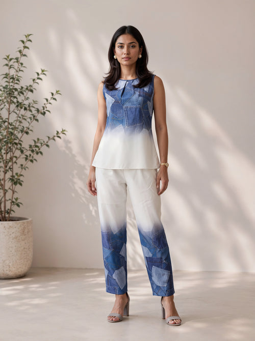 A woman stands in a bright indoor setting wearing a sleeveless blue-and-white gradient top and matching gradient pants with denim-like geometric paneling. She is posed in front of a light beige wall with soft shadows, with a potted plant on the left.