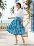 Woman in a white blouse and blue patterned skirt standing in front of a stone building with flowers and palm trees.