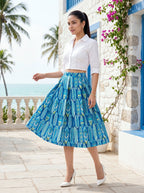 Woman in a white blouse and blue patterned skirt standing in front of a stone building with flowers and palm trees.