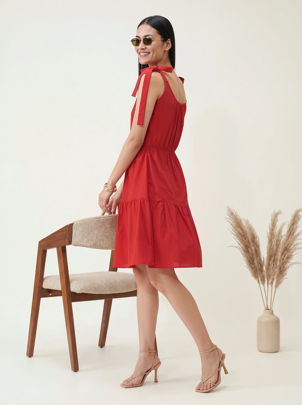 Woman in a red dress standing next to a chair and vase with dried plants on a white background