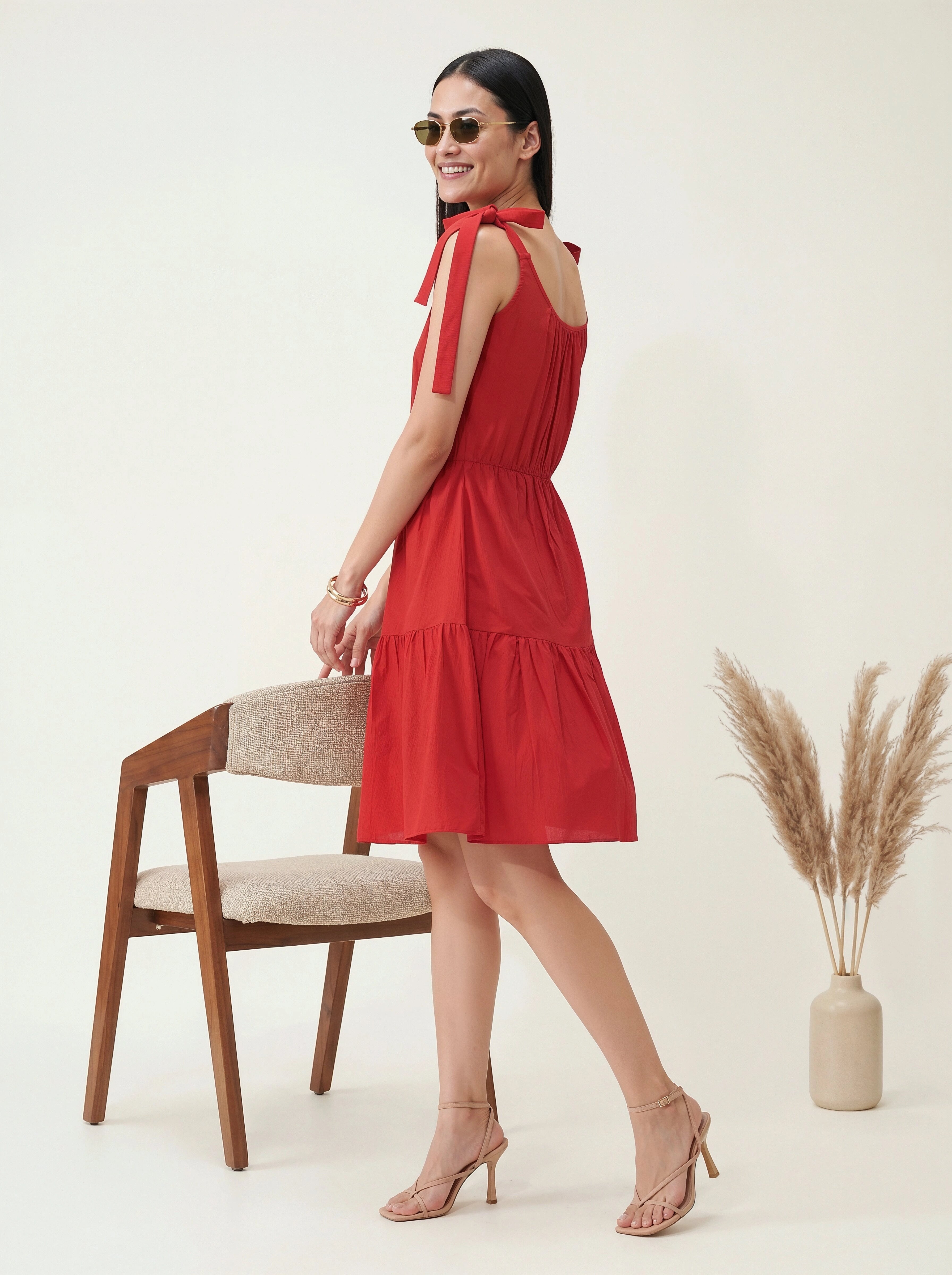 Woman in a red dress standing next to a chair and vase with dried plants on a white background
