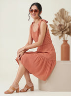Woman in a rust-colored dress sitting on a white surface with a vase and dried plants in the background.