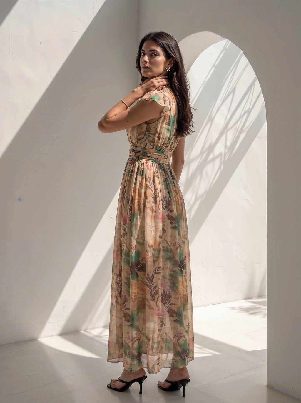 A woman wearing a sleeveless maxi dress with a botanical leaf pattern in beige, green, and brown tones, standing in a minimalist white room with dramatic window shadows.