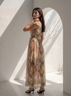 A woman wearing a sleeveless maxi dress with a botanical leaf pattern in beige, green, and brown tones, standing in a minimalist white room with dramatic window shadows.