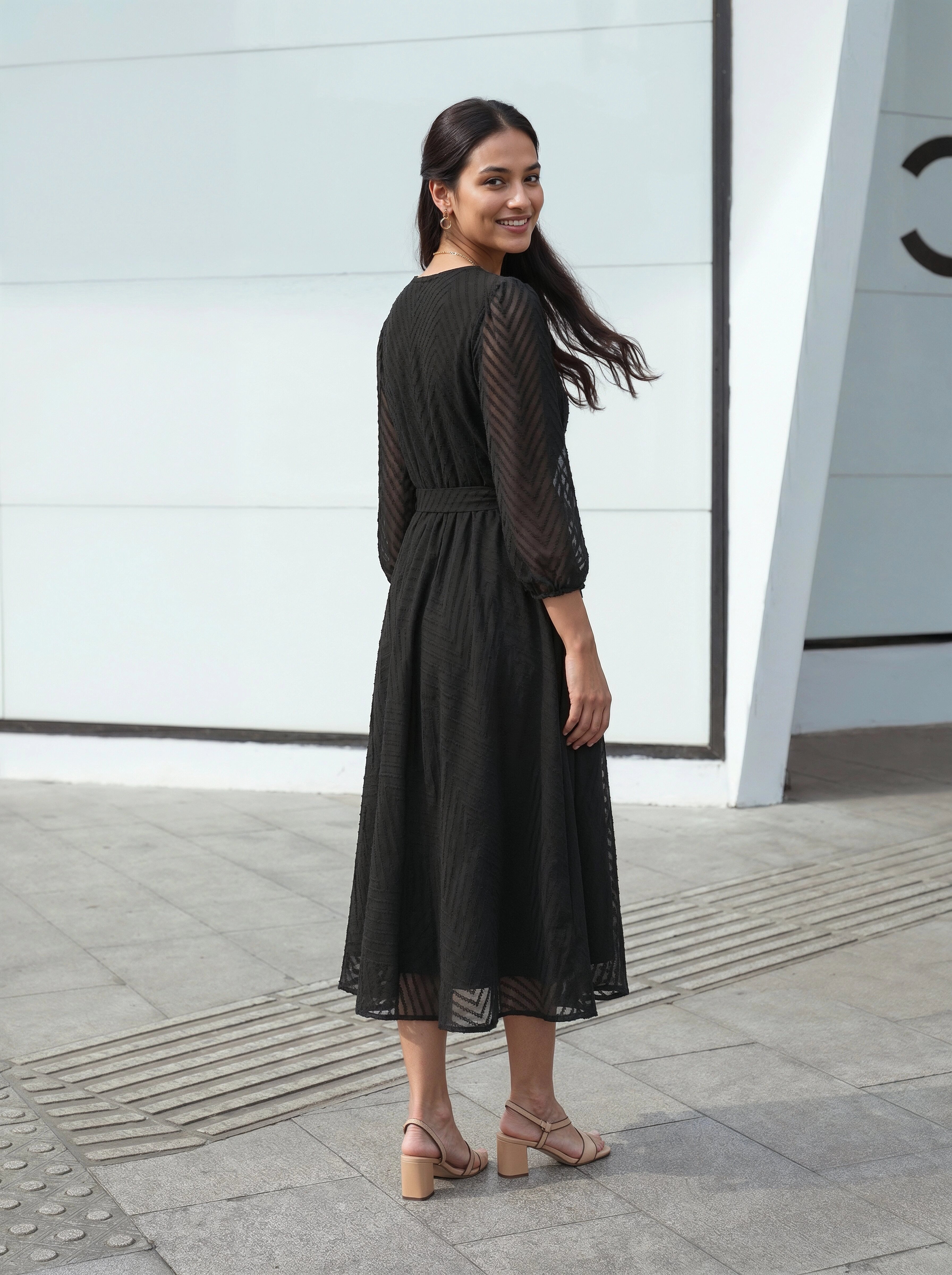 Woman wearing a black dress standing on a sidewalk with a light gray wall in the background