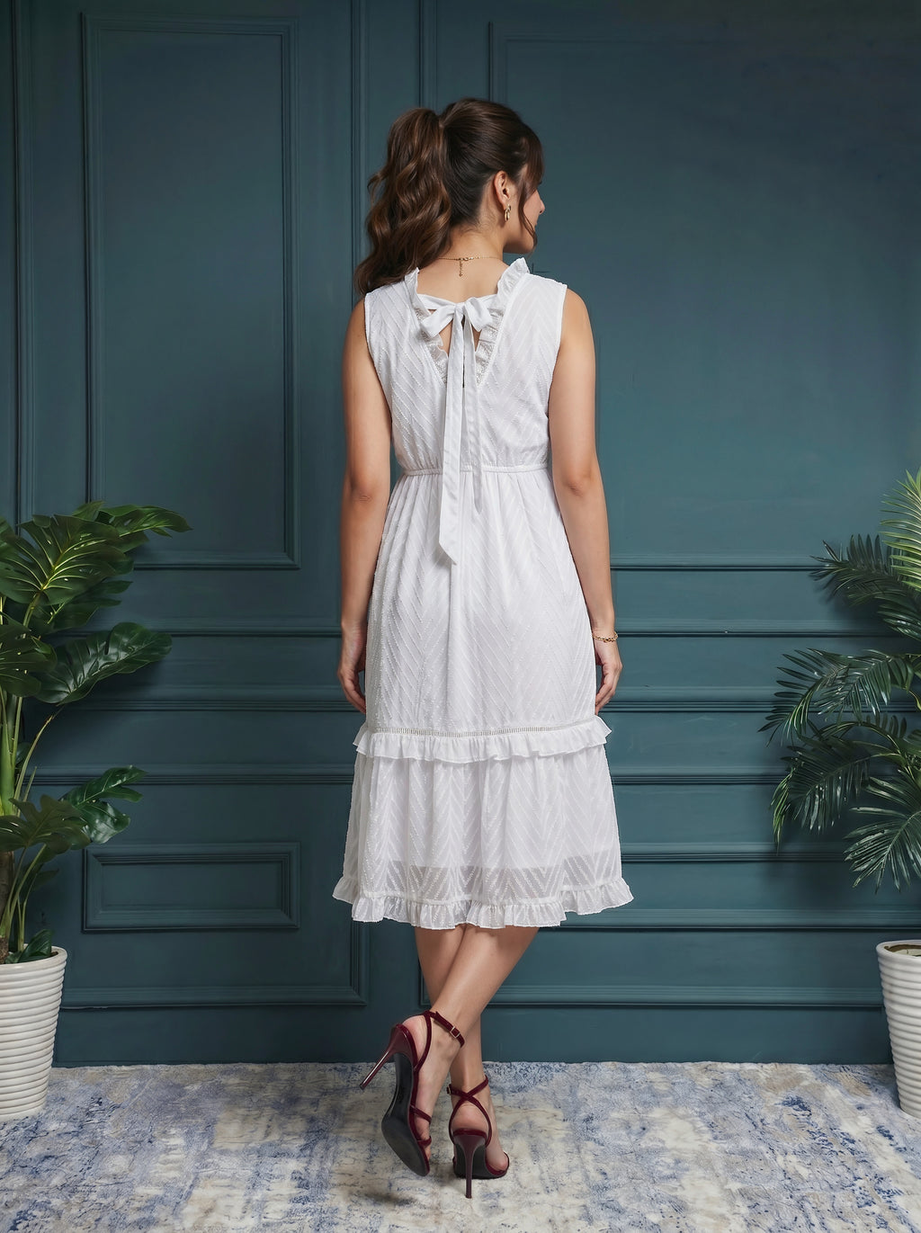 Woman in a white dress standing against a dark blue paneled wall with plants on either side.