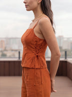 A woman wearing a matching rust-orange two-piece set consisting of a lace-up camisole top and coordinating pants, photographed outdoors on a balcony with a blurred cityscape background.