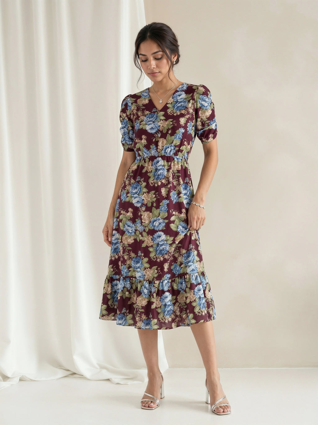A woman models a short-sleeve midi dress with a floral print in burgundy, blue, and beige, standing against a neutral cream backdrop with a draped white curtain.