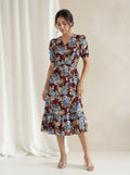 A woman models a short-sleeve midi dress with a floral print in burgundy, blue, and beige, standing against a neutral cream backdrop with a draped white curtain.