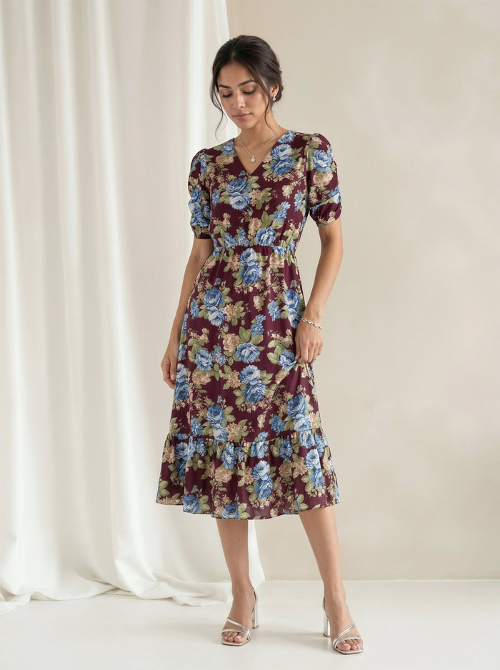 A woman models a short-sleeve midi dress with a floral print in burgundy, blue, and beige, standing against a neutral cream backdrop with a draped white curtain.