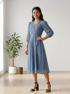 Woman wearing a blue dress standing in a bright room with a plant and wooden stool.