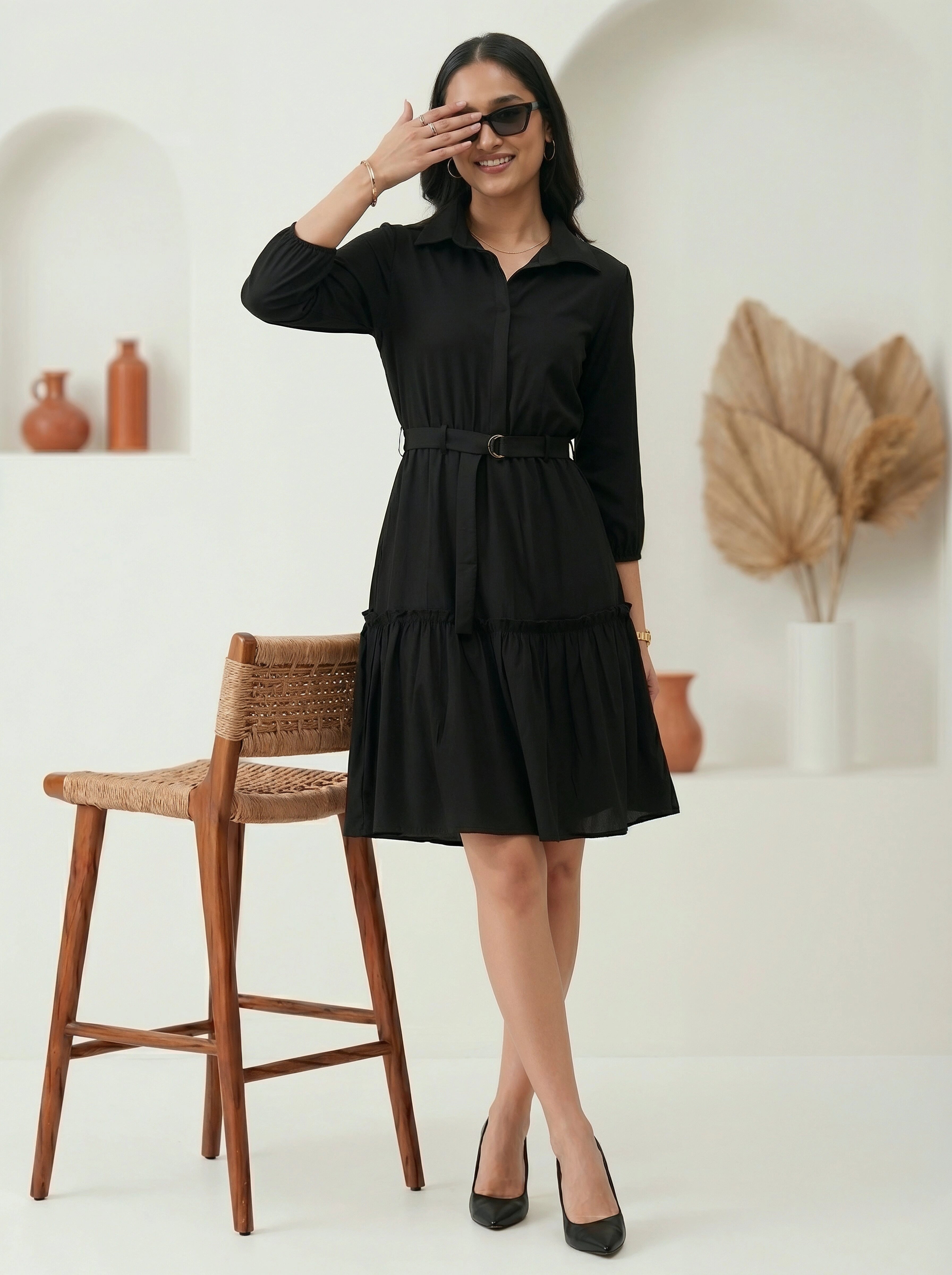 Woman wearing a black dress standing in a room with a wooden chair and decorative items.